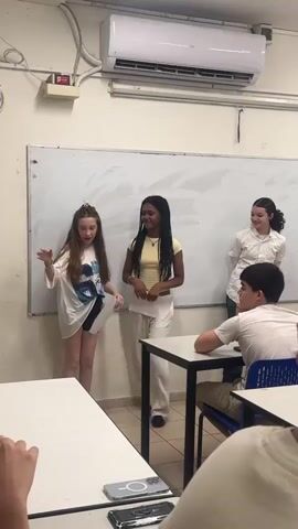 A classroom scene shows three young women standing in front of a whiteboard. The woman on the left, with long brown hair, is wearing a white oversized t-shirt and dark shorts. She is bending down to pick up papers from the floor. The woman in the center, with long dark braided hair, is wearing a yellow crop top and white pants. She is holding papers and looking towards the woman on the left. The woman on the right, with dark curly hair, is wearing a white button-down shirt and jeans. She is also holding papers. In the foreground, two students are seated at desks, with their backs to the camera. The room has a whiteboard, an air conditioning unit, and fluorescent lighting.