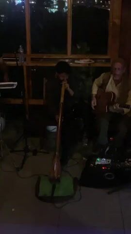 The video captures an indoor musical performance at night, likely in Dharamkot, India. Two men are featured prominently. On the left, a man with dark hair and a beard is seated, playing a didgeridoo, which stands vertically on a green mat on the floor. To his right, another man with long, light-colored hair and a beard is seated, playing an acoustic guitar. Several electronic pedals and cables are on the floor near the guitarist. In the background, large wooden-framed windows offer a view of a dimly lit outdoor scene with scattered lights, suggesting a town or settlement in a hilly region. Wooden beams and shelves are part of the indoor structure. A keyboard stand is partially visible on the far left. The scene depicts a live music event with a relaxed ambiance. No visible text is present.