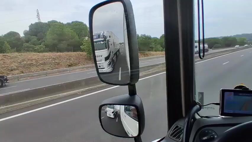This is a first-person perspective video from inside a truck cabin. The camera is positioned to show the driver's view of the road and the side mirrors. The truck is on a highway, driving alongside other vehicles. In the main side mirror, another truck is visible in the adjacent lane, and further down the road, more traffic can be seen. The smaller, lower mirror reflects the back of the truck's own trailer. A gray car passes by in the lane to the left. The environment appears to be a rural highway with trees and greenery along the sides. A GPS device is mounted on the dashboard. The video captures the truck in motion, navigating traffic on the highway.