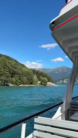 The video pans across a turquoise body of water with waves. A white boat with a red stripe along the top edge is partially visible on the right side of the frame, with a white bench and railing on its deck. The left side of the frame shows a lush green mountainside covered in pine trees. A small, sandy shoreline is visible with a few scattered objects that could be beach umbrellas or tents. The sky is a bright blue with scattered white clouds. In the distance, more mountains are visible, some with snow-capped peaks. The overall scene is a scenic view of a lake or fjord with surrounding mountains and a boat.