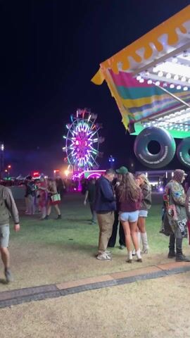 The scene is an outdoor event at night. In the background, a brightly lit Ferris wheel dominates the view. People are milling about in the field. In the foreground, a man with a mustache and a cap walks past the camera. A group of four people stand and talk near a colorful concession stand. The overall atmosphere seems lively and festive.