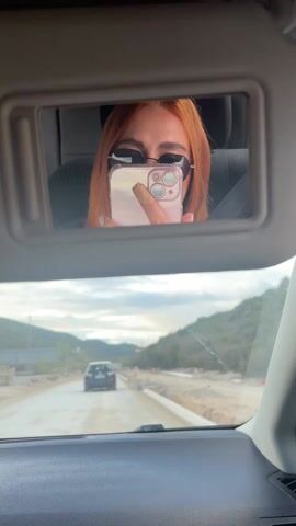 A person with orange hair is driving a car and taking a selfie in the sun visor mirror. The driver is wearing sunglasses. A car is visible in the distance on the road. Construction is occurring on the side of the road. The day is overcast.