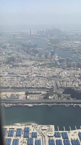 The video presents an aerial view of Dubai from an airplane, with the camera capturing the cityscape, waterways, and transportation infrastructure below. In the foreground, there are a lot of gray buildings, roads, and bridges. In the distance, the Dubai skyline is visible, with several skyscrapers including the Burj Khalifa. The wing of the airplane, emblazoned with the Emirates logo, is briefly visible on the left side of the screen as the camera pans across the landscape. There is a notable amount of haze in the air.