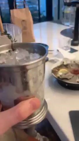 A person holds a metal cup full of ice, a copper-colored straw, and a sprig of greenery. The camera pans to show oysters on a plate with lemon wedges and a small bowl of sauce. The camera continues panning to show the bar, where two bartenders wearing dark shirts are working. A wooden box with compartments is on the bar. The background shows windows looking out onto a cityscape in the evening.