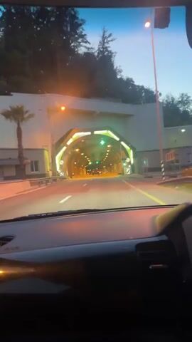 This media file depicts a view from inside a vehicle as it approaches a brightly lit tunnel. The tunnel entrance is framed by an architectural structure with a distinctive, angular design, illuminated by green and yellow lights. Inside the tunnel, a series of lights are visible, creating a glowing pathway. Traffic lights are also present, with some showing green. The road leading into the tunnel appears to be paved, with lane markings visible.

The scene is set in what appears to be a twilight or early evening time of day, as indicated by the darkening sky and the artificial illumination of the tunnel and surrounding structures. Trees and foliage are visible on the hillside above the tunnel entrance, suggesting a natural or semi-natural environment. The overall mood is one of transition, as the vehicle is about to enter a confined, artificial space from the open air. There is no visible text in the media file. The location context provided is Razdolnoye, Russia.