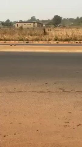The video shows a roadside scene on a somewhat hazy day. The foreground consists of a paved shoulder and a grey asphalt road. Beyond the road is a concrete barrier, followed by a patch of dry grass and scrub. In the background, there's a building constructed of brick or concrete blocks. Some trees and other vegetation are visible in the distance. There's no discernible activity or people visible in the short clip.