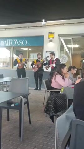 The video shows a group of three male musicians standing and performing in front of a restaurant. They wear traditional costumes with black pants, jackets, and colored sashes. Two of them play guitars, while the third appears to sing.  They stand on the sidewalk under a covered patio area, with the restaurant's entrance behind them.  Several people are seated at tables on the patio, including four young women. They are casually dressed, and appear to be eating. There are dark gray tables and chairs on the patio. The restaurant is called "Arroceria Alfonso". The lighting is bright and the atmosphere seems relaxed.
