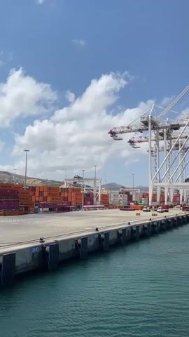 The video shows a large container ship docked at a port. Several large cranes are positioned over the ship, suggesting that cargo is being loaded or unloaded. The port area is filled with stacks of shipping containers of various colors. The water in the harbor is a deep turquoise, and the sky above is blue with scattered white clouds. The overall scene is one of industrial activity at a busy seaport.