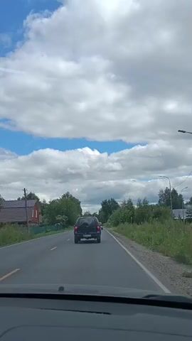 The video shows a view from inside a car driving on a paved road. The sky is blue with scattered white clouds. On the left side of the road, there is a red house with a dark roof, surrounded by trees and a green fence. A utility pole is visible next to the house. Further down the road, a dark SUV is driving away from the camera. On the right side of the road, there are tall grasses and trees. The road has yellow lines marking the lanes. Another car, a dark sedan, is visible ahead of the SUV. The scene appears to be in a rural or suburban area.