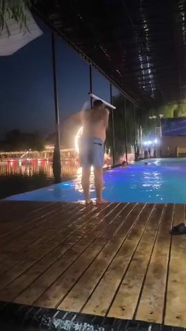 A person jumps into a swimming pool at night. The pool is lit with colorful lights. There is a wooden deck around the pool. In the background, there are more lights and trees. The person is wearing blue shorts. They are not visible after entering the water.