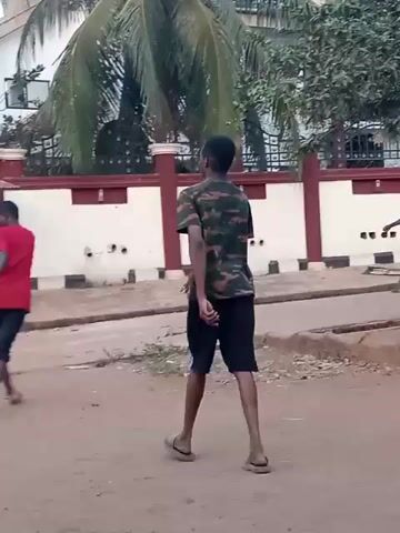 Three young men are playing a game with a ball on a dirt road. The first youth is bent over near the ground. The other two are upright. The first is wearing a red shirt and dark shorts, the other a camouflage shirt and black shorts. A third person is seen briefly. The players interact physically. The setting appears to be a street in a developing area. There are buildings and trees in the background. The day seems bright.