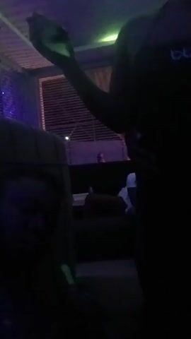 The video shows a dimly lit room with a person in a black dress standing and talking. They appear to be holding something in their hand, possibly a phone, and there is a cloud of smoke in front of them. Other people are seated around the room, and the lighting is provided by blue and purple lights. There's a window with blinds behind the standing person. The overall atmosphere appears to be casual and social, possibly in a lounge or a nightclub setting.