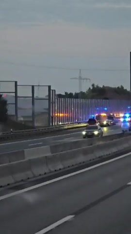 The video shows a highway at dusk or dawn. There is a line of emergency vehicles with flashing blue and red lights on the right side of the highway. These include what appear to be fire trucks and possibly police cars. There are also several other vehicles, including cars and trucks, stopped or moving slowly on the highway. Some of the vehicles appear to have their hoods open. There is smoke or steam rising from the road surface in a few places. The weather appears to be overcast. The scene suggests a traffic incident or accident.