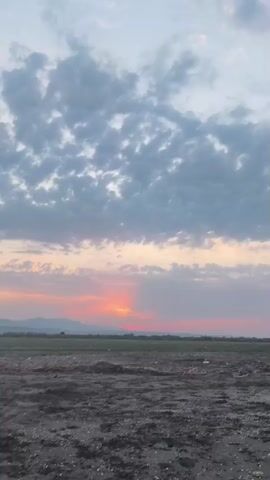 The video pans across a landscape at dusk. In the foreground, there is a patch of rough, dark earth with some debris scattered on it. In the midground, a flat plain stretches out, dotted with some sparse vegetation and possibly a few distant trees. In the background, a mountain range is visible, silhouetted against the sky. The sky itself is filled with a mixture of pale blue and soft gray clouds, with the sun setting on the horizon, casting a warm orange and pink glow. The light from the setting sun creates a visible beam or shaft of light extending upwards through the clouds. The overall mood is peaceful and serene. No people or animals are visible in the video.