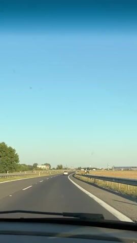 A car is driving down a highway. The sky is clear and blue. On the left side of the road, there are trees and a guardrail. On the right side of the road, there is a guardrail and fields. The road is paved and has white lines marking the lanes. Another car is visible in the distance.