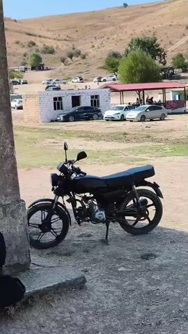 A black motorcycle is parked in the foreground. In the background, there is a dirt road, a small building with a white exterior and a red-tiled roof, and several cars parked. Further in the background, there are hills with sparse vegetation under a clear blue sky. A person is partially visible in the lower left corner of the frame.