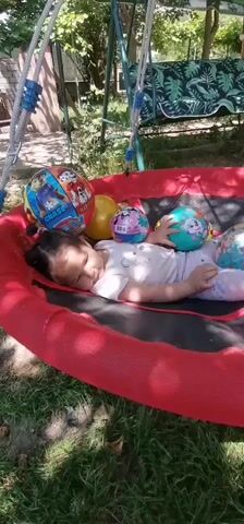 In this media file, a young child is playing on a saucer swing with several balls. The child, appearing to be a toddler, is lying on the red fabric of the swing, surrounded by a variety of colorful balls, some decorated with characters. The swing is suspended from a metal frame, and the background suggests an outdoor setting with trees, grass, and part of a patio visible. A bench swing with a patterned cushion is also in the background. The lighting indicates it is daytime, likely sunny, casting shadows on the ground. The child’s expression is content, suggesting enjoyment of the activity. There is no visible text in this media file. The setting appears to be a residential backyard, possibly in Osoitsa, Bulgaria, as indicated by the provided location context.