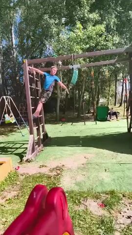 A young male is climbing on a wooden playground structure with a ladder and horizontal bars. He is wearing a blue t-shirt and patterned shorts. He is holding onto one of the horizontal bars with his right hand and has his left leg on the ladder. He appears to be mid-climb or in the process of transitioning. The playground has green artificial turf and is surrounded by trees. In the background, another person is visible sitting on a green slide, and a swing set is also present. A red object, possibly part of another play structure, is in the foreground, partially obscuring the view. The weather appears to be sunny.