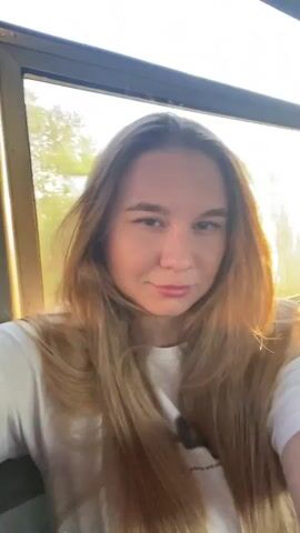 The media file is a selfie-style video of a young woman with long, blonde hair, wearing a white t-shirt. She is sitting indoors, likely in a vehicle given the motion blur of the scenery outside the window. The lighting suggests it is late afternoon or early evening, with the sun casting a warm glow. The woman's expression shifts from a neutral gaze to a slight smile, then she turns her head to look out the window before returning to face the camera with a subtle smirk. The background outside the window shows trees and greenery, indicating a rural or semi-rural setting. There is no discernible text visible in the video. The overall mood is casual and relaxed, possibly capturing a moment during a journey.