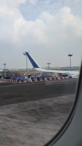 A view from an airplane window shows the wing of an airplane. The plane is on an airport tarmac. In the background, there are cargo containers and airport lights. The sky is partly cloudy.