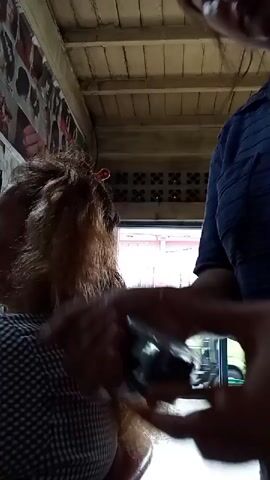 The video shows a close-up of a person's hands holding a clear plastic bottle with a metallic cap and a brown substance inside. The person is about to pour the substance from the bottle into their palm. In the background, another person's head and shoulders are visible, with long, reddish-brown hair. They are wearing a checkered shirt. The environment appears to be outdoors, possibly a patio or covered area, with a corrugated metal roof overhead and a brick wall in the background. The lighting suggests it is daytime.