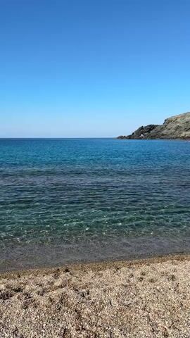 A vertical video shows a calm, clear blue sea with a rocky coastline visible in the distance on the right. The foreground consists of a pebble beach. The sky is a cloudless, bright blue. The water is transparent enough to see the pebbles on the seabed in the shallow area near the shore, transitioning to a deeper turquoise color further out. The overall impression is one of a serene, sunny coastal landscape.