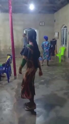 The video shows an indoor scene with a woman in a colorful dress standing with her back to the camera. She is wearing a dress that is patterned with shades of green, red, and brown, and has a ruffled black piece over her shoulders. She has long black hair down her back and brown shoes. In the background, there are other people sitting in chairs, standing, and milling about. Several blue plastic chairs are visible, along with a green plastic chair. A red pole is centrally positioned in the room. The room itself has simple, painted walls, a window with bars, and bare floors. A framed picture hangs on the wall. The lighting is provided by overhead bulbs, which are somewhat glaring. Music can be heard.