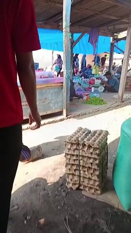 A person wearing a red shirt and black pants walks past stacks of egg cartons in what appears to be an outdoor market. In the background, other people are visible, some sitting and some standing, near stalls with various goods. There are also some motorcycles and structures covered with tarps. The ground is dusty and uneven.