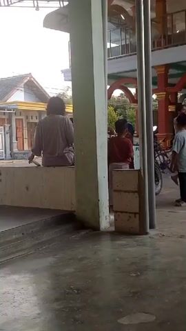 A group of people are standing and sitting near a building with an arched entrance. A bicycle and a scooter are parked nearby. One person is walking away from the camera. The ground is paved with concrete.