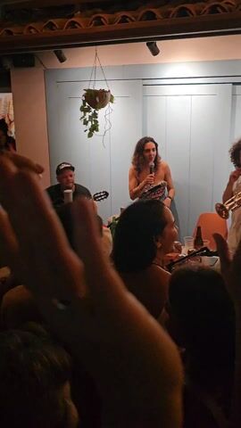 A music band consisting of a female singer, a guitarist, and a trumpeter performs in front of a live audience in what appears to be a small, cozy bar or a house. The band stands on a makeshift stage, under soft lighting. The singer, wearing a patterned top and shorts, holds a microphone and sings animatedly. The guitarist, wearing a hat, plays a guitar. The trumpeter, sporting curly hair and a patterned shirt, plays the trumpet while smiling. The audience is closely packed together, watching and enjoying the performance. Some people in the crowd are moving, likely dancing to the music. The room has a low ceiling with a rustic design, with wooden beams and what seems to be traditional roof tiles. Hanging plants add a touch of greenery to the scene. The atmosphere is lively and intimate.