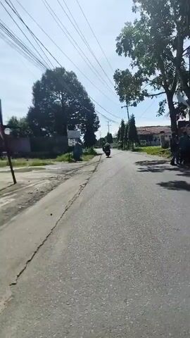The video shows a first-person perspective of someone riding a scooter down a paved road. The rider is looking ahead, and the scooter's dashboard is visible in the foreground. The road is lined with trees and some houses. There are power lines overhead. In the distance, other motorcycles can be seen. The sun is shining, and the sky is blue. There are tire marks visible on the road. The overall environment appears to be a rural or suburban area.