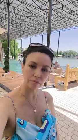 A woman with dark hair is posing for a selfie. She is wearing sunglasses on her head and a light blue dress with an abstract pattern. Behind her, there is a swimming pool with several people in it, a wooden deck, and a body of water with boats in the distance. The scene appears to be outdoors with a covered structure overhead.