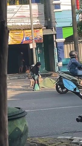 A white car is parked on the right side of the frame. A motorcycle with two people wearing red helmets passes by. A woman wearing a black headscarf and a patterned dress walks across the street with a plastic bag. The scene appears to be a street in a town with buildings and shops in the background.