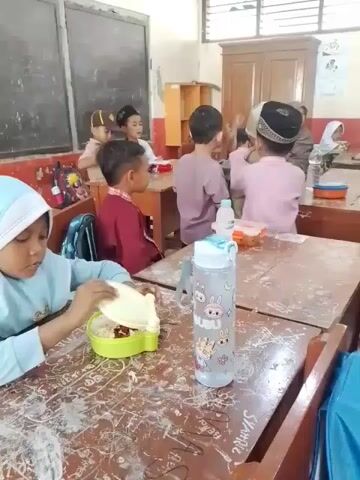 The video shows a classroom with several young children seated at desks, seemingly during lunchtime. A girl in a light blue hijab sits at a desk in the foreground, opening a green lunchbox. A water bottle with cartoon designs is placed next to the lunchbox. Other children are seated at desks in the background, some eating, others talking amongst themselves. Some boys wear traditional Muslim caps. The desks are wooden and appear aged with scribbles on the surfaces. There is a blackboard on the wall, a wooden cabinet, and windows that allow natural light into the room. The atmosphere appears lively with children engaging in conversation and eating.