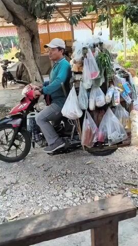 A man in a light blue shirt, gray pants, and a white cap sits on a red motorcycle loaded with various goods in plastic bags. The motorcycle is parked on a gravelly surface near a tree and a makeshift wooden stall. The man is looking towards the camera and appears to be speaking. The goods on the motorcycle include packaged snacks, bottles, and what look like fresh produce. The environment suggests a rural or outdoor market setting.