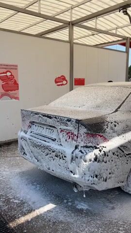 A person is using a hose to spray soapy water onto a black car. The car is covered in thick white foam, indicating it is being washed. The scene takes place at an outdoor car wash bay with a covered roof. The background shows white walls, red accents, and some trees. The car has a license plate with the numbers 1772 FGQ. The person is wearing a white shirt and glasses. The action appears to be the initial sudsing stage of washing the car.