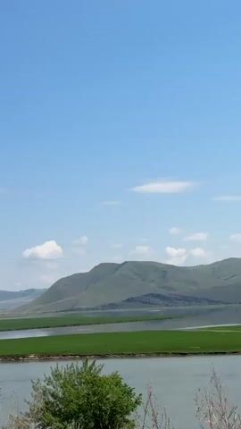 The video shows a landscape view with a body of water in the foreground and rolling green hills in the background. The sky is clear and blue with a few scattered clouds. The camera pans slowly across the scene, capturing the vastness of the landscape. There are no people or objects clearly visible in the video, except for what appears to be a car's side mirror in the bottom right corner of the frame, reflecting a person's arm and hand. The environment appears to be natural and undeveloped.