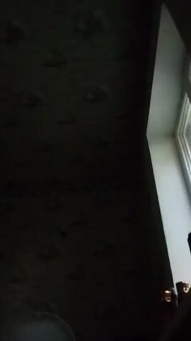The video shows a close-up view of a windowsill in a dimly lit room. Light streams in from a window on the right side of the frame, illuminating several objects on the windowsill. From left to right, there are several red-capped bottles, a black cylindrical object, and a purple cylindrical object. The background on the left is dark, with what appears to be patterned wallpaper. The angle of the camera is looking upwards towards the window.