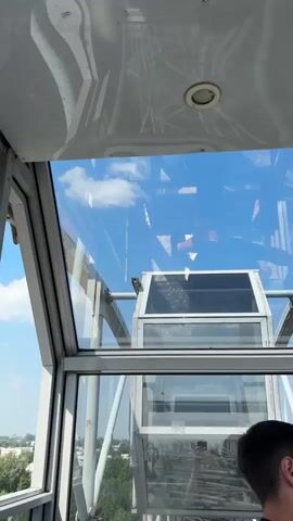 A man in the foreground looks to his right, his right ear and side of his head visible. Behind him, the view from inside a glass enclosed structure, possibly a Ferris wheel car, shows a clear blue sky with a few white clouds. The structure appears to be made of metal and glass. Through the glass, a distant industrial building with a tall brick structure and conveyors can be seen, surrounded by green trees and some smaller buildings. A red-bordered sign with text is affixed to the glass. The camera pans slightly, revealing more of the surrounding landscape.