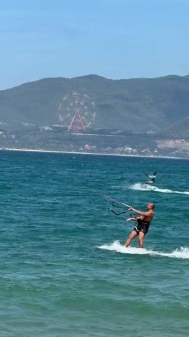 A man kitesurfs on the ocean. He is on a board with a kite attached to him. He is wearing black shorts. The water is blue and wavy. In the background, there are green hills and a Ferris wheel. The sky is clear and blue. There is another kitesurfer in the distance.