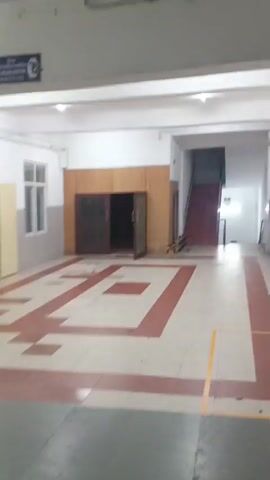 The video shows an indoor scene, possibly a hallway or common area. The floor is tiled with a geometric pattern of red and cream squares. There are wooden cabinets with open doors in the center of the frame. To the right, a staircase with red steps leads upwards. Windows are visible on the left wall. In the second clip, a rolling cart with shelves is seen. The third clip shows a closed dark door with signs on the wall above it, and a red arrow painted on the floor pointing to the right. The fourth clip shows a dimly lit area with patterned flooring. The final clip shows a long hallway with the same patterned flooring and fluorescent lighting. There are no people visible in the video.