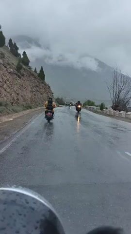 The video shows a group of motorcyclists riding on a wet, paved road in a mountainous area. The sky is overcast and the weather appears to be rainy. Trees and a rocky slope are visible on the left side of the road. Mountains covered in clouds are in the background. One motorcyclist in the foreground is partially visible, showing part of their motorcycle and the road ahead. Several other motorcycles are visible further down the road, also moving in the same direction. One motorcyclist is wearing a yellow jacket and a backpack. The road is reflective, indicating it is wet.