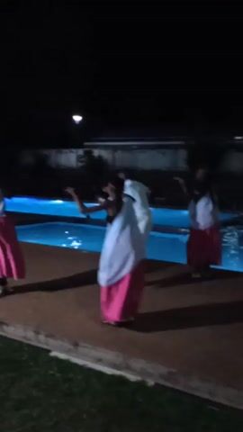 In a nighttime video, four people dance beside an illuminated swimming pool. Three individuals wear pink skirts and white tops, while the fourth wears a white outfit. They are dancing on a concrete patio with grass visible at the edge. The background includes a building and streetlights. The pool emits a bright blue glow against the darkness.