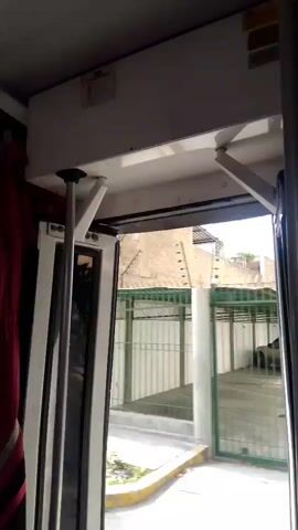 The video shows the interior of what appears to be a bus or a similar vehicle. The camera is positioned near the entrance, looking outwards. The door is open, revealing a view of the outside environment. The outside consists of a paved area with a fence and a building in the background. The fence appears to be made of metal poles and mesh. There is a car visible parked behind the fence. The overall lighting suggests it is daytime.