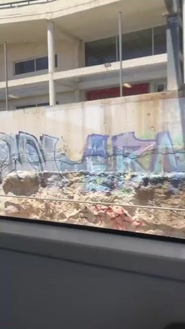 The media file captures a dynamic scene from inside a vehicle, looking outwards towards a concrete wall covered in vibrant graffiti. The wall appears to be part of an elevated structure, possibly a highway overpass or a retaining wall, with a multi-story building visible in the background. This building has a light-colored facade, several rows of windows, and a repeating pattern of vertical support columns. The weather appears to be sunny, with clear skies, suggesting daytime.

The primary focus of the media is the extensive graffiti artwork adorning the concrete wall. The graffiti features a variety of tags and pieces in different colors, including silver, black, green, blue, and purple. The style of the graffiti is typical of urban street art, with bold lettering and abstract designs. Some of the graffiti includes what appears to be stylized lettering or signatures.

Interspersed with the graffiti are a few signs. One sign is green and yellow with a pictogram of a person running, possibly an emergency exit or safety instruction sign. Another sign is a road marker showing "Km. 6" and "Fim", which translates to "Kilometer 6" and "End" in English and Catalan respectively, providing a location cue.

In the background, beyond the graffiti wall, cars are visible traveling on a road or highway. The vehicles are moving, indicating ongoing traffic. The building behind the wall also shows glimpses of activity, with what appear to be more cars in a parking area or on a lower level.

The perspective suggests the media was recorded by a passenger or driver in a moving vehicle, likely a car, as evidenced by the window frame and interior elements at the bottom of the frame. The overall impression is one of an urban or semi-urban environment, possibly on the outskirts of a city, characterized by infrastructure, traffic, and street art. The time of day is clearly daytime due to the bright sunlight and shadows. No people are distinctly visible or identifiable in the media.