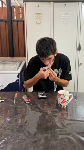A young man with dark hair and a black t-shirt is sitting at a table with a dark, possibly plastic, tablecloth. He is holding a small silver object in his hands and appears to be working on it. In front of him on the table, there is a mobile phone, a patterned mug, and a knife. To his left, a white and blue cooler is visible, and behind him, there are white lockers. The lighting suggests an indoor environment, possibly a communal area. The man's focus is on the object in his hands, and he appears to be manipulating it with precision.
