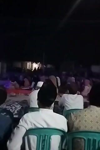 The video shows a nighttime gathering of people, likely outdoors, with some illumination from lights. In the foreground, several individuals are seated on green plastic chairs, facing away from the camera and towards a larger group of people. These foreground individuals are dressed in white clothing and some wear head coverings. The larger group in the background appears to be seated on the ground and on chairs, with many individuals facing a central area, suggesting a presentation, performance, or religious gathering. The environment is dark with some ambient light, hinting at a rural or semi-rural setting.