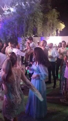 The video shows a group of people dancing at what appears to be an outdoor evening event, possibly a wedding reception. The lighting is dim and colorful, with purple and blue hues prominent. Several people are visible in the foreground and background, some dancing energetically, others watching. One woman in a white, lacy dress is in the center foreground, her back to the camera, her arms extended. To her right, a woman in a blue dress is also dancing. A man in a white shirt is in the background, holding a child on his shoulders. The environment appears to be a garden or patio area with trees and possibly a bar in the distance. The overall atmosphere seems festive and celebratory.