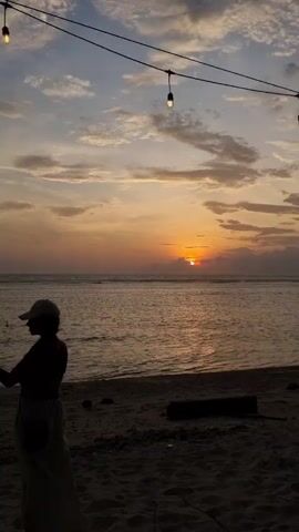The video shows a woman taking a selfie at sunset on a beach. The sky is orange and blue with clouds. A string of lights hangs above. The woman is silhouetted against the sunset and wearing a hat.