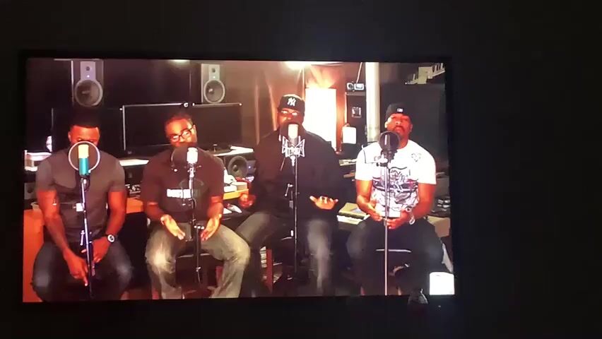 The video displays a medium shot of four Black men in a recording studio. They are seated in front of microphones, suggesting a musical performance or recording session. The men are casually dressed, with one wearing a New York Yankees baseball cap. The studio is equipped with professional audio gear, including monitors and mixing boards, indicating a professional setting. The lighting is warm and intimate, creating a focused atmosphere. The men appear engaged and focused on their performance. The overall mood is serious and concentrated, typical of a creative recording environment. There is no visible text within the frame. The time of day and weather are not discernible from the image. The location is given as Angeles, Philippines.
